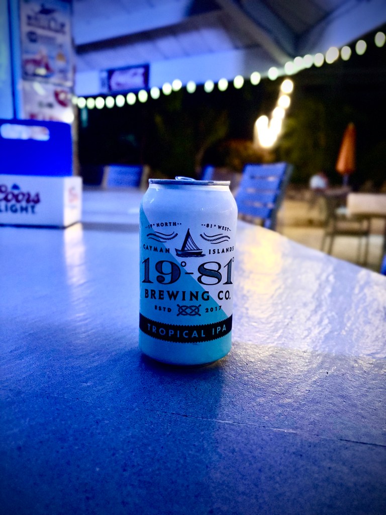 A can of 19°81 Brewing Co. Tropical IPA placed on a table at night, with a softly lit background featuring blue and green hues.